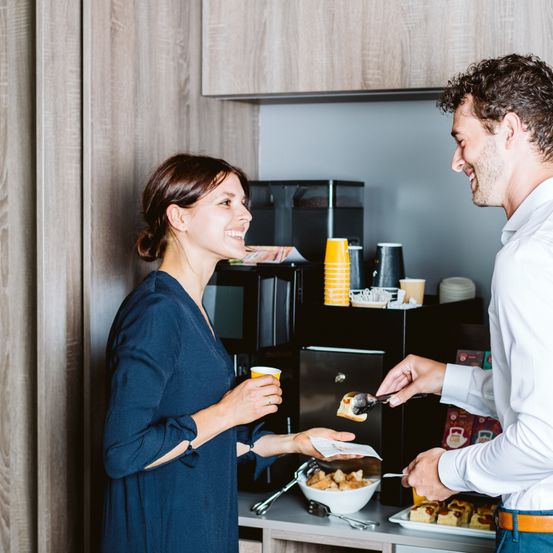 Ein Mann und eine Frau stehen lächelnd in einer Küche. Der Mann hält Essen mit einer Zange, während die Frau einen Becher hält. Hinter ihnen steht eine Kaffeemaschine und eine Schüssel mit Essen auf dem Tresen.