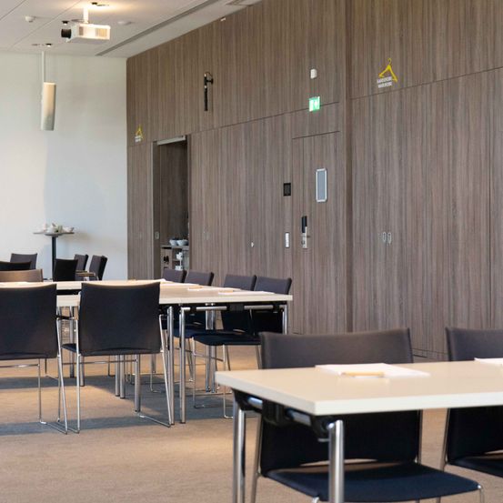 Ein leerer Konferenzraum mit einem langen Tisch und Stühlen, Holzwänden und einer offenen Tür. Der Raum ist gut beleuchtet, und es gibt mehrere kleine Tische an der Seite.