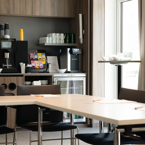 Ein gut ausgestatteter Büro-Pausenraum mit moderner Einrichtung. Tische mit Stiften, einer Kaffeemaschine und einem kleinen Kühlschrank. Tassen und Schüsseln ordentlich angeordnet. Ein großes Fenster mit Vorhang.