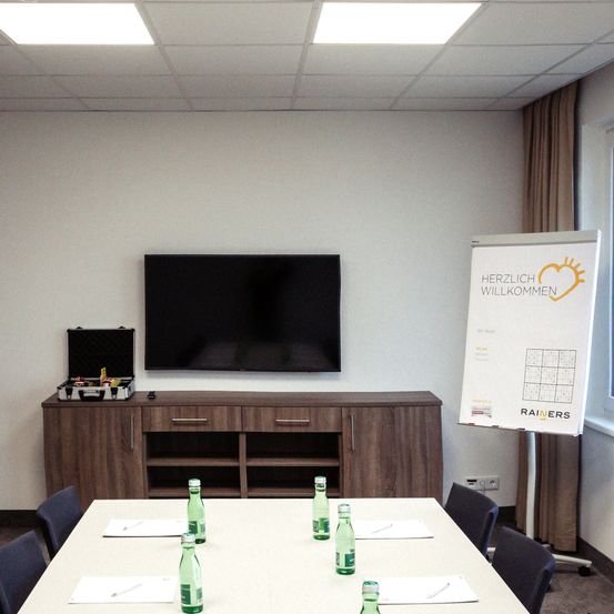 Ein Besprechungsraum mit einem Schreibtisch, Stühlen und einem Fernseher an der Wand. Auf dem Schreibtisch steht eine Aktentasche und an der Wand hängt eine Tafel mit einem Rätsel.