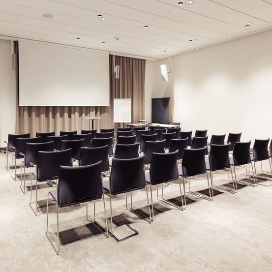 Ein leerer Konferenzraum mit Reihen von Stühlen vor einer großen Projektionsleinwand, einem Podium mit einer Tafel und Deckenleuchten.