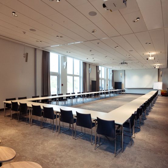 Ein großer Konferenzraum mit weißen Wänden und einem langen Tisch, umgeben von Stühlen. Es gibt große Fenster mit Vorhängen, eine Projektionsleinwand und Deckenleuchten.