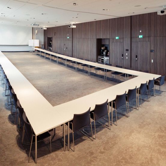 Ein Konferenzraum mit langen Tischen und Stühlen ist leer. Der Raum hat einen großen, gebogenen Tisch mit vielen Stühlen, die darum angeordnet sind. Der Raum hat Holzwände und einen Teppichboden.