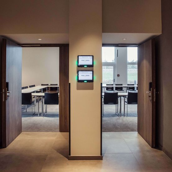 Ein Konferenzraum mit leeren Tischen und Stühlen, einer weißen Wand und zwei Monitoren an einem Pfeiler. Es gibt offene Türen auf beiden Seiten.