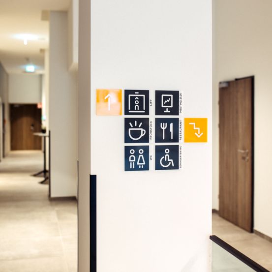 Ein Innenkorridor mit einer weißen Wand, die verschiedene Bodenschilder enthält, einschließlich Aufzug, Besprechungsraum, Rezeption, Cafeteria, Restaurant und Behinderteneinrichtungen.
