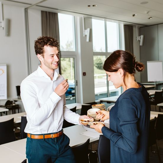 Ein Mann in einem weißen Hemd und blauer Hose steht in einem Klassenzimmer, lächelt und unterhält sich mit einer Frau in einem blauen Hemd.