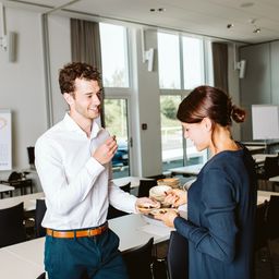 Ein Mann in einem weißen Hemd und blauer Hose steht in einem Klassenzimmer, lächelt und unterhält sich mit einer Frau in einem blauen Hemd.