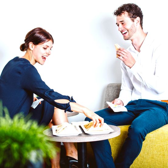 Eine Frau und ein Mann sitzen auf einem Sofa und essen von Tellern. Der Mann lächelt und die Frau schaut ihn an. Sie sind in einer Innenumgebung.