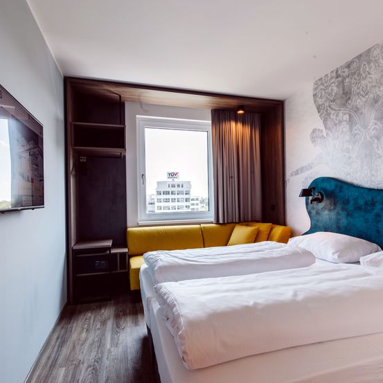 Ein Hotelzimmer mit zwei Betten, einem gelben Sofa, einem Fenster mit Stadtblick, einer Lampe und einem Fernseher an der Wand.