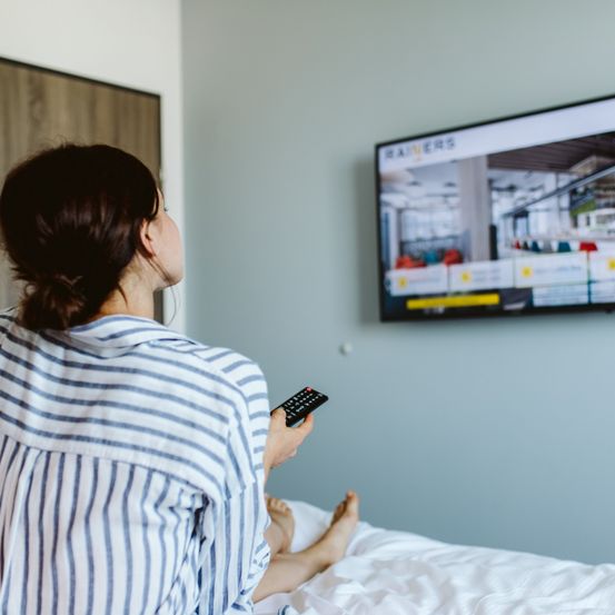 Eine Frau in einem gestreiften Shirt sitzt auf einem Bett und schaut fern. Der Fernsehbildschirm zeigt ein Bild einer Bibliothek.