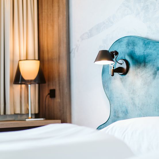 Ein Schlafzimmer mit einem blauen Kopfteil und weißer Bettwäsche, einer Lampe auf dem Nachttisch und einer wandmontierten Lampe. Der Raum hat einen Holzkopfteil und eine weiße Wand mit einem Design.