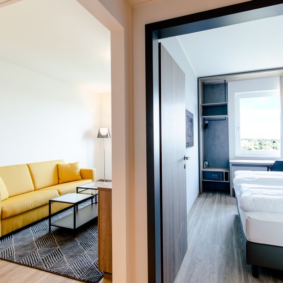 Ein modernes Hotelzimmer mit einem gelben Sofa, einem Couchtisch und einem Bett mit weißen Laken. Das Zimmer verfügt über ein großes Fenster, einen Kleiderschrank und eine Lampe.