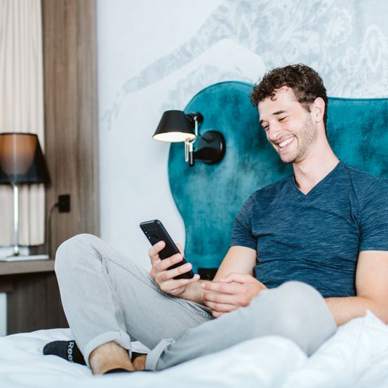Ein lächelnder Mann sitzt auf einem Bett und hält ein Handy. Er trägt ein blaues T-Shirt und graue Hosen. Eine Lampe steht auf dem Nachttisch.