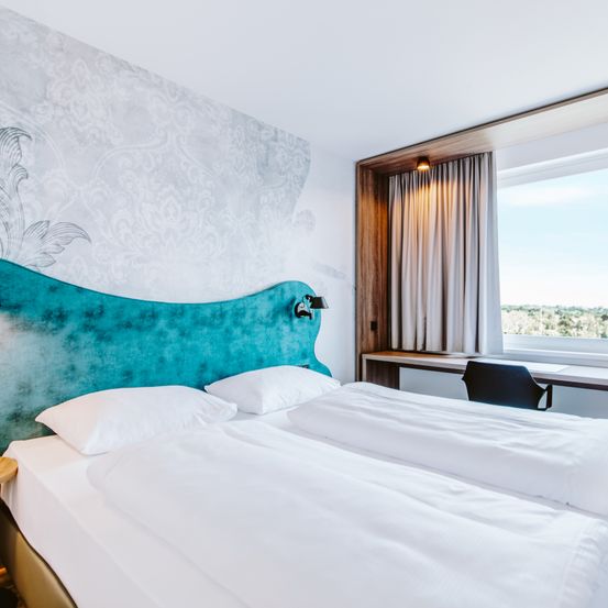 Ein Zimmer mit zwei Betten, weißer Bettwäsche und einem blauen Kopfteil mit Wanddekoration. Ein Fenster mit Vorhängen und einem Schreibtisch mit schwarzem Stuhl.