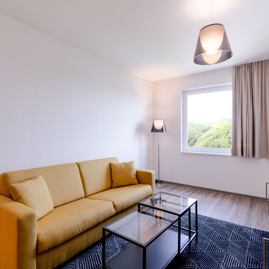 Ein modernes Wohnzimmer mit einem gelben Sofa, Glastisch und einem großen Fenster mit Blick aufs Freie. Der Raum hat weiße Wände, einen Holzboden und dekorative Lampen.