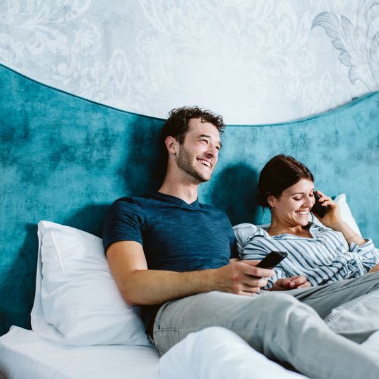 Ein lächelndes Paar sitzt auf einem Bett, ein Mann hält ein Handy und eine Frau telefoniert. Sie schauen beide in die Kamera und genießen ihre Zeit.