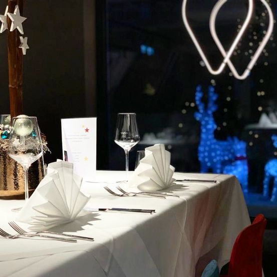 Ein Tisch für eine Mahlzeit gedeckt mit Weingläsern, gefalteten Servietten und Besteck. Im Hintergrund sind ein Weihnachtsbaum, ein Stern und leuchtende Rentiere.