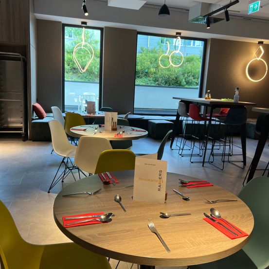 Ein moderner Essbereich mit mehreren runden Tischen, jeder mit Besteck, Menüs und dekorativen Lichtern. Die Umgebung umfasst verschiedene farbige Stühle und Sofas, mit großen Fenstern, die Ausblicke aufs Freie bieten.