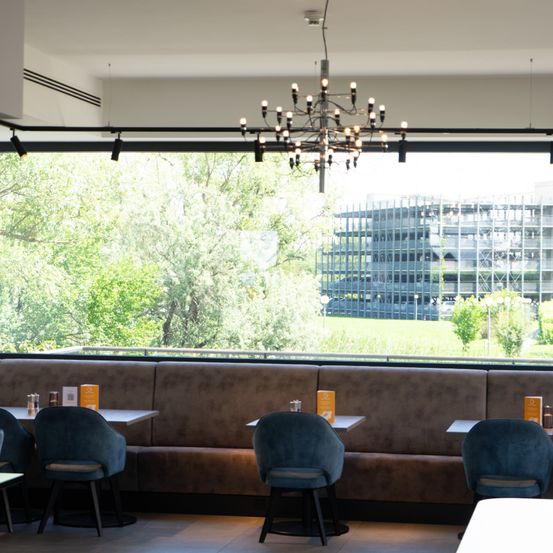 Ein Restaurant mit einem großen Fenster mit Blick auf ein Feld und ein Gebäude im Bau. Im Inneren gibt es eine Kristallleuchte und Stühle an Tischen.