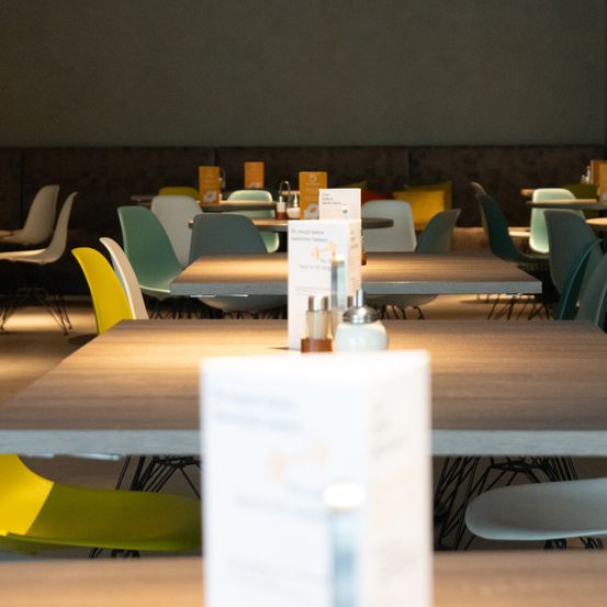 Ein Restaurant mit modernem Design bietet eine Vielzahl von farbenfrohen Stühlen um Tische herum an. Die Tische sind mit Gewürzen und Menükarten ausgestattet, was auf einen Essbereich hinweist. Die Beleuchtung ist weich und schafft eine gemütliche Atmosphäre.