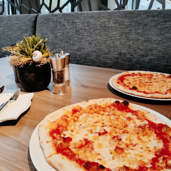 Zwei Pizzen mit geschmolzenem Käse auf weißen Tellern stehen auf einem Holztisch. Ein Topf mit Pflanzen und Gewürzmühlen sind ebenfalls auf dem Tisch. Ein grauer gepolsterter Stuhl steht hinter dem Tisch.