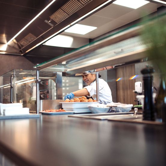 Eine Köchin bereitet in der Küche Essen zu und trägt Handschuhe. Tabletts und Schüsseln stehen auf dem Tresen.