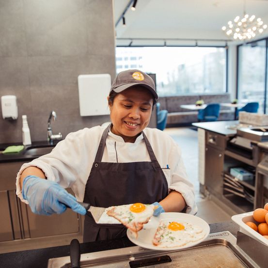 Eine Frau mit Schürze lächelt und hält eine Pfanne mit zwei gebratenen Eiern auf einem Teller.