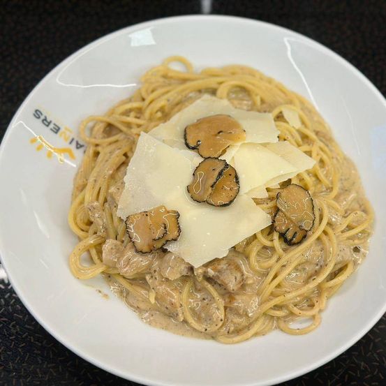 Ein Teller mit Nudeln in cremiger Sauce, überbacken mit Käse und Pilzscheiben, auf einem weißen Teller serviert. Der Teller hat das Logo eines Restaurants.