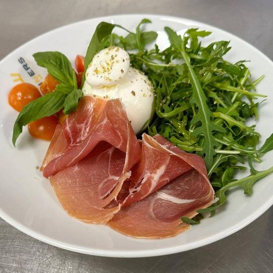 Ein weißer Teller zeigt ein Gericht mit Prosciutto-Scheiben, einer Burrata-Kugel, Kirschtomaten und Rucolablättern.
