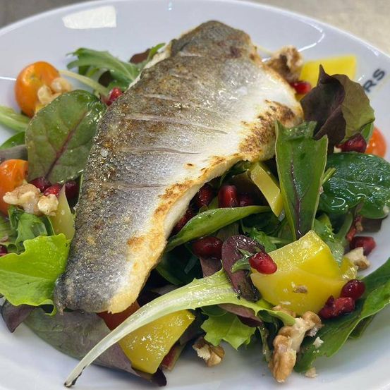 Ein Teller mit frischem Salat, darauf ein ganzer Fisch, garniert mit Kirschtomaten, Granatapfelsamen und Walnüssen. Der Fisch ist leicht gebräunt.