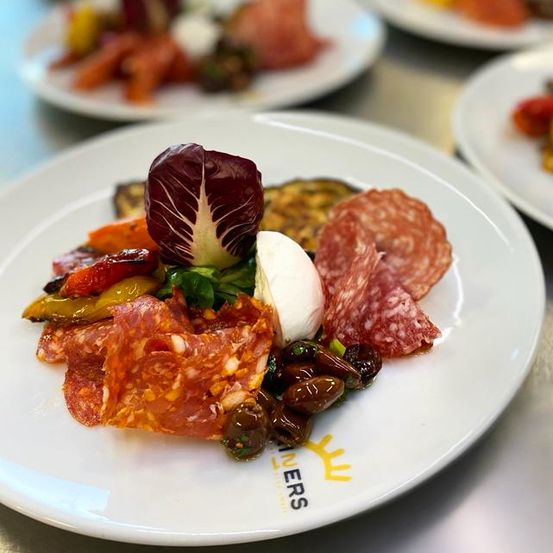 Ein weißer Teller mit verschiedenen Wurstwaren, Gemüse und Käse. Das Gericht hat roten Kohl, Tomaten und Oliven. Der Teller ist mit dem Wort 'Wineers' gebrandet.