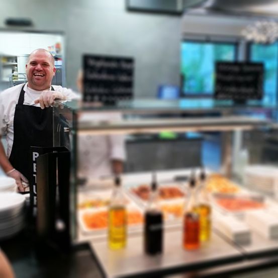 Ein Koch steht hinter einem Buffettisch in einem Restaurant. Er trägt eine schwarze Schürze mit 'Petit' darauf. Flaschen mit Saucen sind ausgestellt. Am unteren linken Rand ist die Hand einer Person zu sehen.