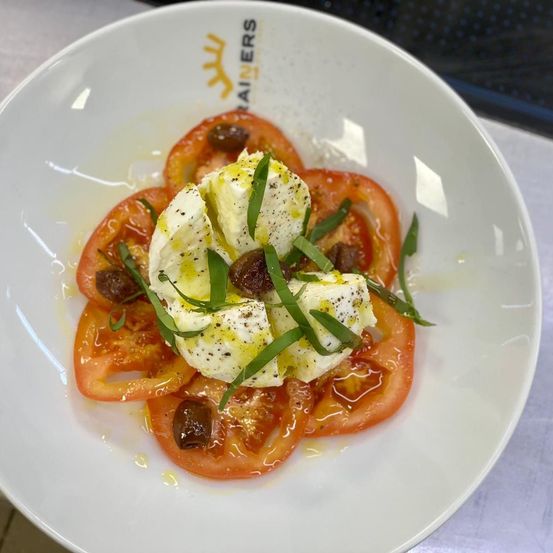 Ein Teller mit Tomatenscheiben, belegt mit Käse und Kräutern. Das Gericht hat einen leichten Olivenölguss. Der Teller steht auf einer grauen Oberfläche.