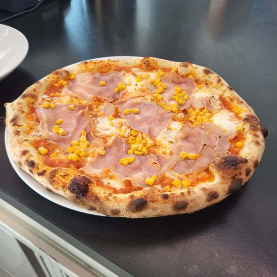 Eine Pizza mit Schinken und Mais auf einem weißen Teller steht auf einer schwarzen Arbeitsplatte mit einem weißen Teller links.