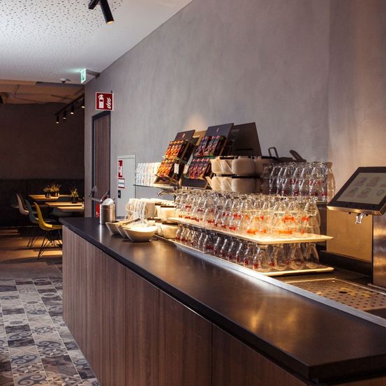 Eine moderne Bartheke mit ordentlich angeordneten Gläsern, Schalen und einem Monitor. Ein Feuerlöscher ist an der Wand dahinter montiert. Im Hintergrund sind Tische und Stühle aufgestellt.