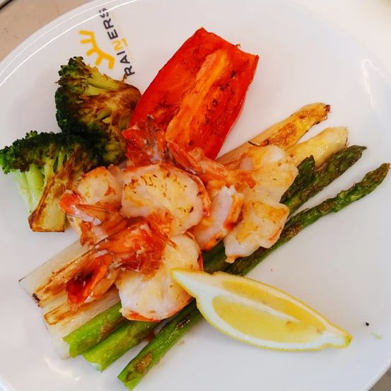 Ein weißer Teller mit Garnelen, Spargel, Brokkoli und einer Zitronenscheibe. Die Garnelen sind gekocht und auf den Spargel und Brokkoli gelegt. Die Zitronenscheibe befindet sich auf der rechten Seite. Der Teller hat ein Markenlogo darauf.