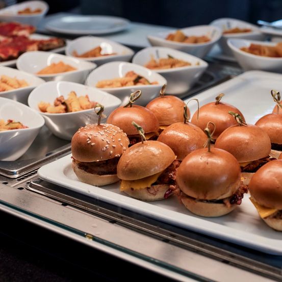 Ein Tablett mit mehreren Mini-Burgern mit Sesam und Zahnstochern, auf einem Tresen mit anderen Gerichten platziert.