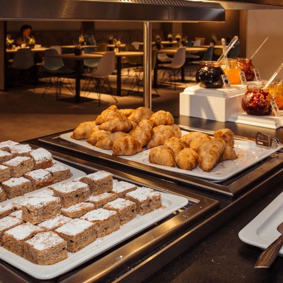 Ein Buffettisch mit Gebäck, einschließlich Croissants und quadratischen Kuchen, steht unter einer Decke mit Deckenbeleuchtung. Der Raum hat Stühle und Tische, und eine Frau sitzt an einem.