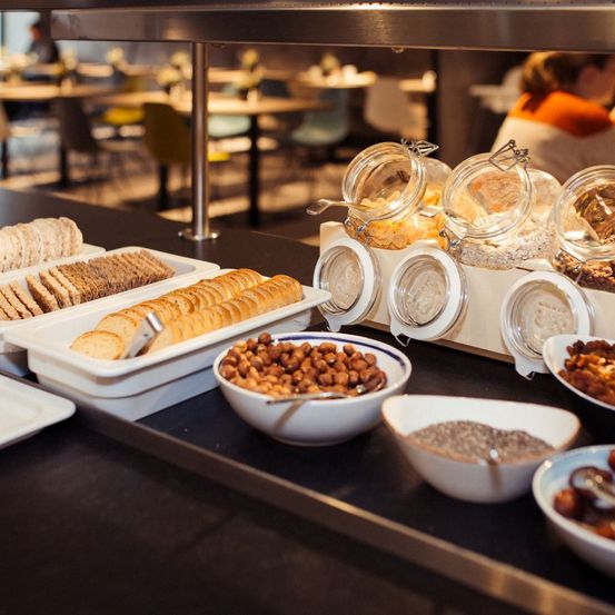 Ein Buffettisch bietet eine Auswahl an Snacks, einschließlich Brot, Nüssen und Trockenfrüchten, die in Gläsern und Schalen präsentiert werden. Die Umgebung scheint ein Essbereich mit Tischen und Stühlen im Hintergrund zu sein.