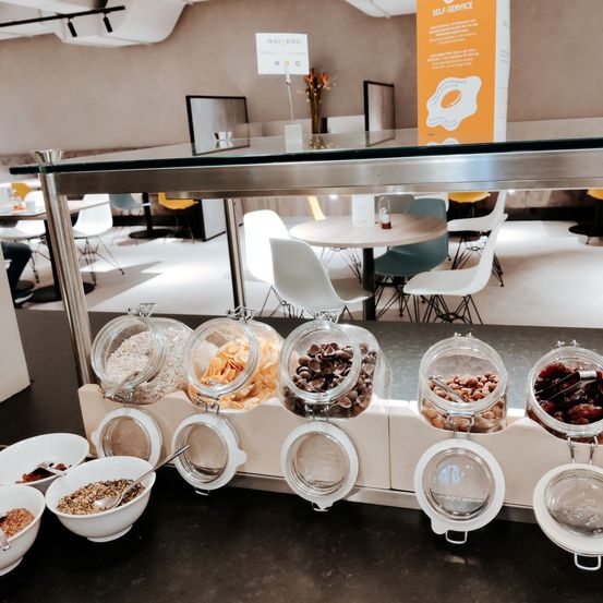 Ein moderner Essbereich mit einem Glasregal, das verschiedene Snacks in durchsichtigen Gläsern anzeigt. Schüsseln mit Essen und ein Löffel sind auf dem Tisch. Ein Schild an einem Pfosten lautet 'SELBSTDIENST'.