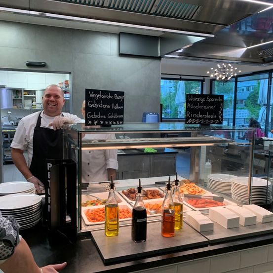 Ein Koch steht hinter einer Vitrine mit verschiedenen Lebensmitteln. Die Tafeln zeigen Optionen wie vegetarische Burger und Käsekrainer an. Stapel von Tellern sind in der Nähe.