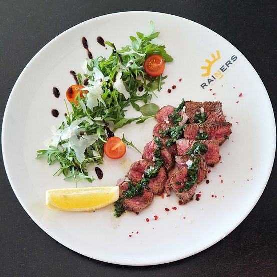 Ein Teller mit Steak, garniert mit Kräutern und Zitrone, begleitet von einem Salat mit Rucola, Tomaten und leichter Vinaigrette. Der Teller trägt das Logo von Rainers.