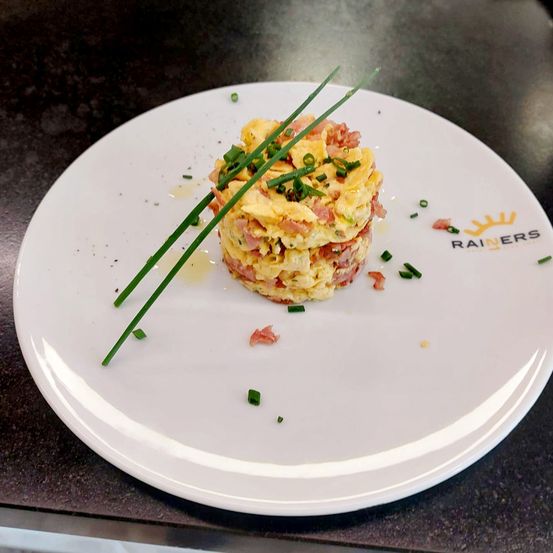 Ein Teller mit einem runden, geschichteten Gericht, gekrönt mit Frühlingszwiebeln, Schinken und einem Spiegelei, präsentiert von Rainers.