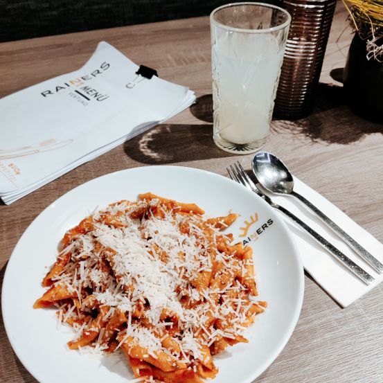 Ein Teller mit Pasta in Tomatensoße und geriebenem Käse steht auf einem Tisch mit Menü, Löffeln und einem Glas Milch.