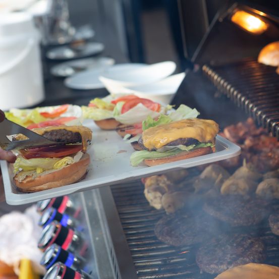 Eine Person schneidet einen Cheeseburger auf einem Tablett, während andere Burger auf einem Grill gegrillt werden.