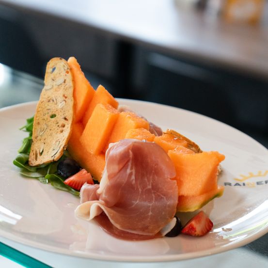 Ein weißer Teller enthält eine Melonenscheibe, eine Prosciutto-Scheiben und eine Brotscheibe mit grünen Blättern. Der Teller hat das Logo 'Rainer' darauf.