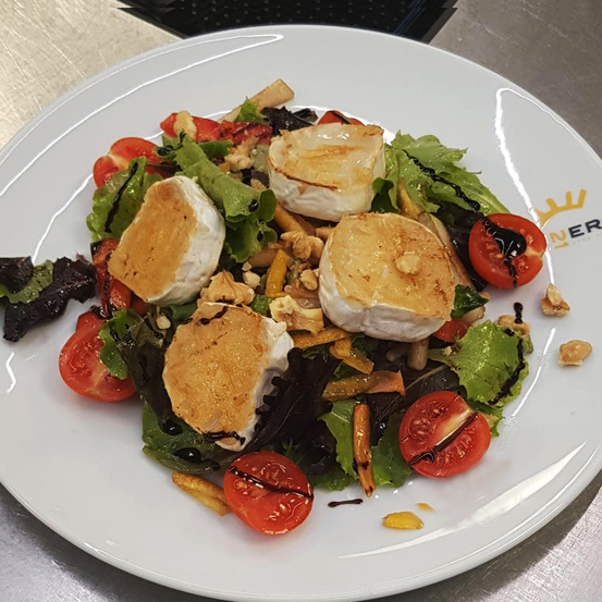 Ein weißer Teller mit einem Salat aus gegrilltem Ziegenkäse, Kirschtomaten und gemischten Blattsalaten, überzogen mit Balsamico-Glasur und mit Nüssen bestreut.