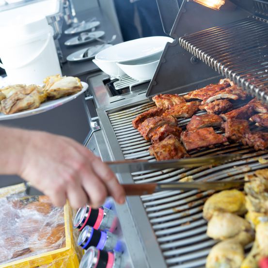 Hände halten Zangen über Rippen auf einem Grill, mit anderem Fleisch und Essen im Hintergrund.