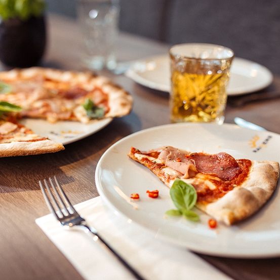 Eine Pizza auf einem Teller mit einem Glas Bier und eine Gabel auf einem Tisch. Ein weiterer Teller mit einer Pizza ist teilweise gegessen.