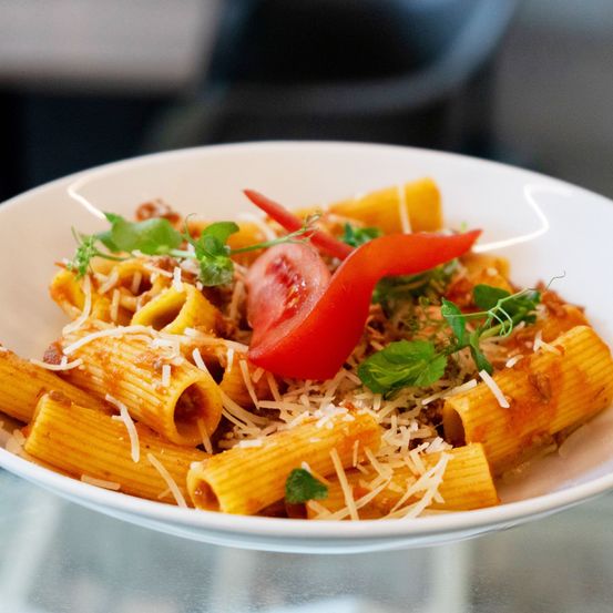 Eine weiße Schüssel mit Rigatoni-Pasta, roter Sauce, Tomatenscheibe und grünen Kräutern obenauf, garniert mit geriebener Käse.